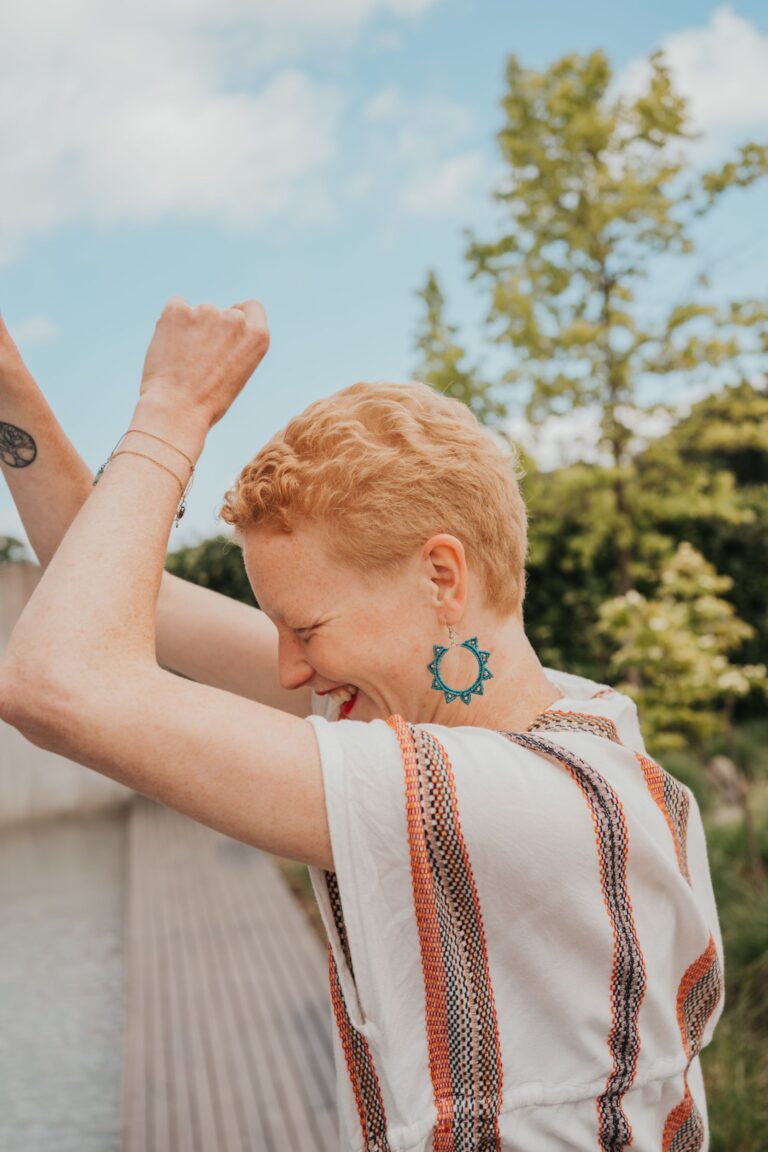 Eine lächelnde Person mit kurzen, lockigen Haaren hebt lachend die Arme in die Luft. Sie trägt auffällige türkisfarbene Ohrringe und ein gestreiftes Oberteil. Im Hintergrund sind Bäume, blauer Himmel und ein Holzsteg am Wasser zu sehen